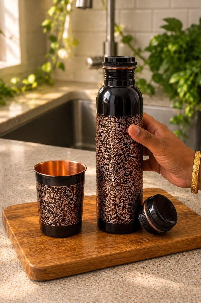K&ouml;nnte beinhalten: Eine schwarze Metallwasserflasche und ein passender Trinkbecher, beide mit einem komplizierten kupferfarbenen Blumenmuster versehen. Die Flasche hat einen abnehmbaren schwarzen Schraubverschluss und das Innere des Bechers zeigt ein poliertes Kupfer-Finish.