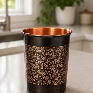 K&ouml;nnte beinhalten: Ein zylindrischer Metallbecher mit einem polierten Kupferinneren und einer dunklen, oxidierten Au&szlig;enfl&auml;che. Der K&ouml;rper ist mit einem komplizierten, ge&auml;tzten Blumenmuster verziert, das die helle Kupferfarbe unter der dunklen Oberfl&auml;che zeigt.