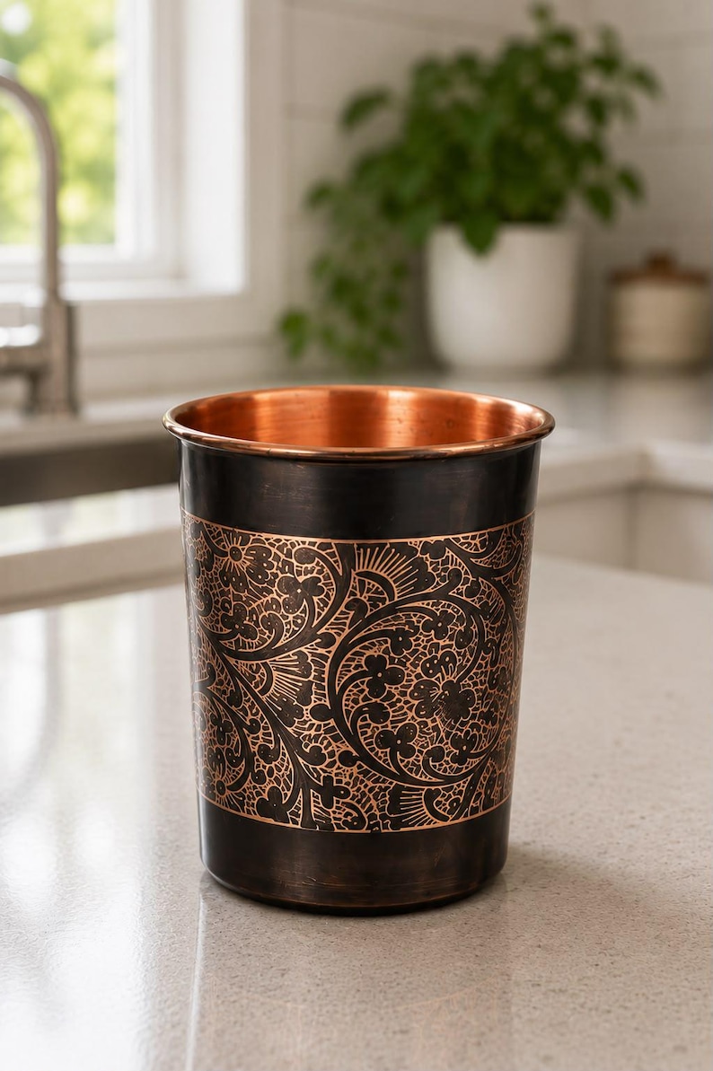 K&ouml;nnte beinhalten: Ein zylindrischer Metallbecher mit einem polierten Kupferinneren und einer dunklen, oxidierten Au&szlig;enfl&auml;che. Der K&ouml;rper ist mit einem komplizierten, ge&auml;tzten Blumenmuster verziert, das die helle Kupferfarbe unter der dunklen Oberfl&auml;che zeigt.