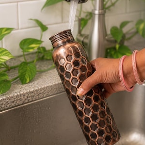 K&ouml;nnte beinhalten: Eine zylindrische Wasserflasche aus Kupfer mit einem erhabenen, sechseckigen Wabenmuster auf der Au&szlig;enfl&auml;che. Die Flasche hat einen schmalen Hals mit einem Gewindeverschluss und eine dunkle, br&uuml;nierte Metalloberfl&auml;che mit hellen Akzenten.