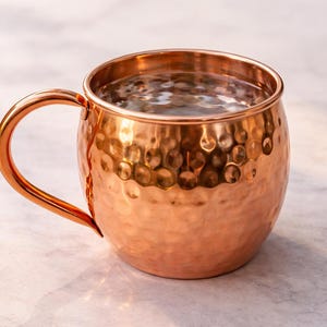May include: A copper Moscow Mule mug with a hammered texture and a curved handle. The mug is filled with a clear liquid, likely a cocktail. The copper has a warm, reddish-brown hue, and the mug sits on a light-colored surface.