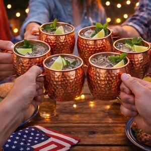 May include: Six copper Moscow Mule mugs filled with ice, lime wedges, and mint sprigs are raised in a toast. The mugs have a hammered texture and are held by people at an outdoor gathering. Food and an American flag are visible.