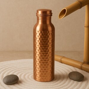 May include: A copper-colored water bottle with a textured, dimpled surface. The bottle is cylindrical with a screw-on cap. The image includes a bamboo water feature and two gray stones, suggesting a spa or zen theme.