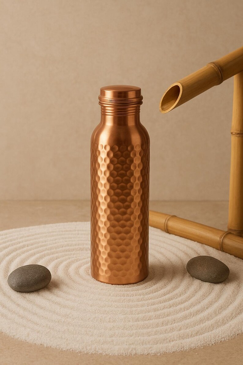 May include: A copper-colored water bottle with a textured, dimpled surface. The bottle is cylindrical with a screw-on cap. The image includes a bamboo water feature and two gray stones, suggesting a spa or zen theme.