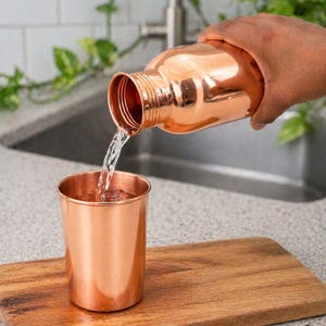 May include: A copper water bottle is pouring water into a copper cup. The cup sits on a wooden cutting board. The bottle and cup have a shiny, metallic finish. The background includes a kitchen sink and greenery.