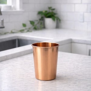 May include: A copper-colored metal cup with a smooth, polished surface. The cup is cylindrical with a slightly flared rim. It sits on a light-colored countertop in a kitchen setting. A plant in a white pot is visible in the background.