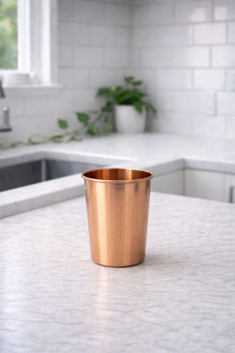 May include: A copper-colored metal cup with a smooth, polished surface. The cup is cylindrical with a slightly flared rim. It sits on a light-colored countertop in a kitchen setting. A plant in a white pot is visible in the background.
