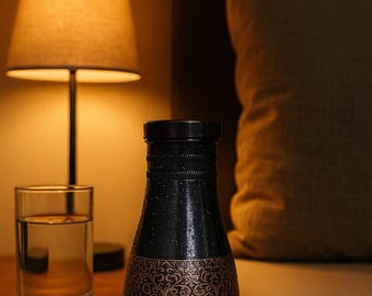 Op de afbeelding: Een zwarte decoratieve fles met een gouden patroon, waarschijnlijk voor wijn of sterke drank, staat op een houten nachtkastje. Een glas water en een lamp met een warme gloed staan ook op de tafel.