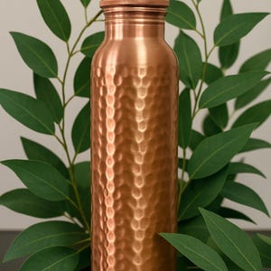 May include: A copper water bottle with a textured, hammered finish. The bottle has a screw-on cap and is surrounded by green leaves. The bottle is approximately 10 inches tall.