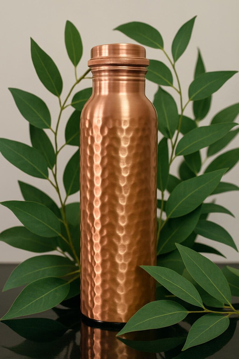 May include: A copper water bottle with a textured, hammered finish. The bottle has a screw-on cap and is surrounded by green leaves. The bottle is approximately 10 inches tall.