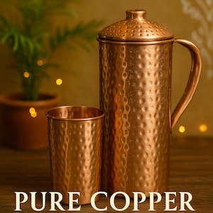 Puede incluir: Una jarra de cobre con un vaso a juego. La jarra tiene tapa y asa. Ambos artículos tienen una textura martillada. El texto de la imagen dice "PURE COPPER, HANDCRAFTED IN INDIA, BOOSTS IMMUNITY."