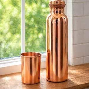 May include: A copper water bottle and matching cup sit on a wooden surface in front of a window. The bottle is tall and cylindrical with a screw-on cap. The cup is short and round, reflecting the warm light.