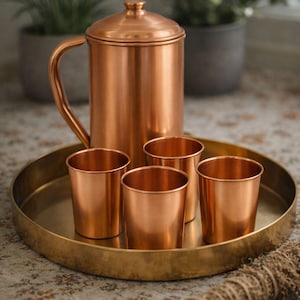 May include: A copper tea set, including a lidded pitcher with a curved handle and four small cups, sits on a round brass tray. The set is displayed on a patterned surface, suggesting a kitchen or dining setting.