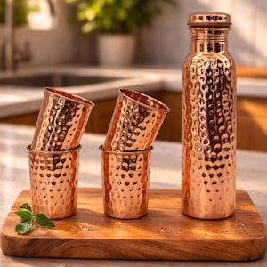 Könnte beinhalten: Eine Kupferwasserflasche und vier passende Becher mit gehämmerten Strukturen werden auf einem Holzschneidebrett präsentiert. Die Flasche ist hoch und zylindrisch mit einem Schraubverschluss. Die Becher sind paarweise gestapelt.
