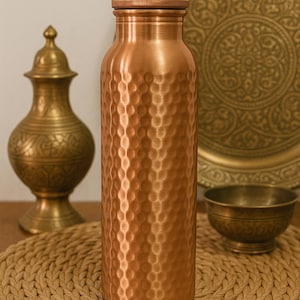 May include: A copper water bottle with a textured, dimpled surface. The bottle has a screw-on cap and a black base. Other copper and brass decorative items are in the background, including a plate, bowl, and vase. The bottle is sitting on a woven placemat.