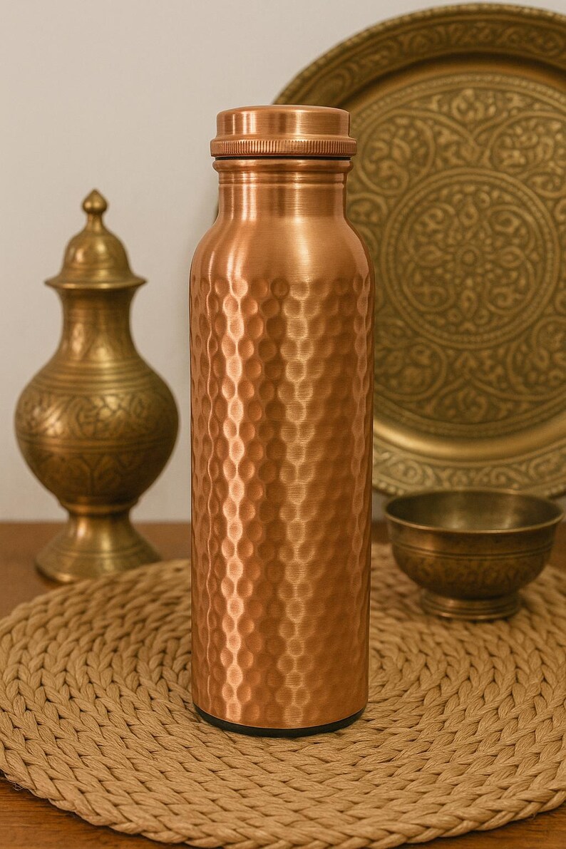 May include: A copper water bottle with a textured, dimpled surface. The bottle has a screw-on cap and a black base. Other copper and brass decorative items are in the background, including a plate, bowl, and vase. The bottle is sitting on a woven placemat.