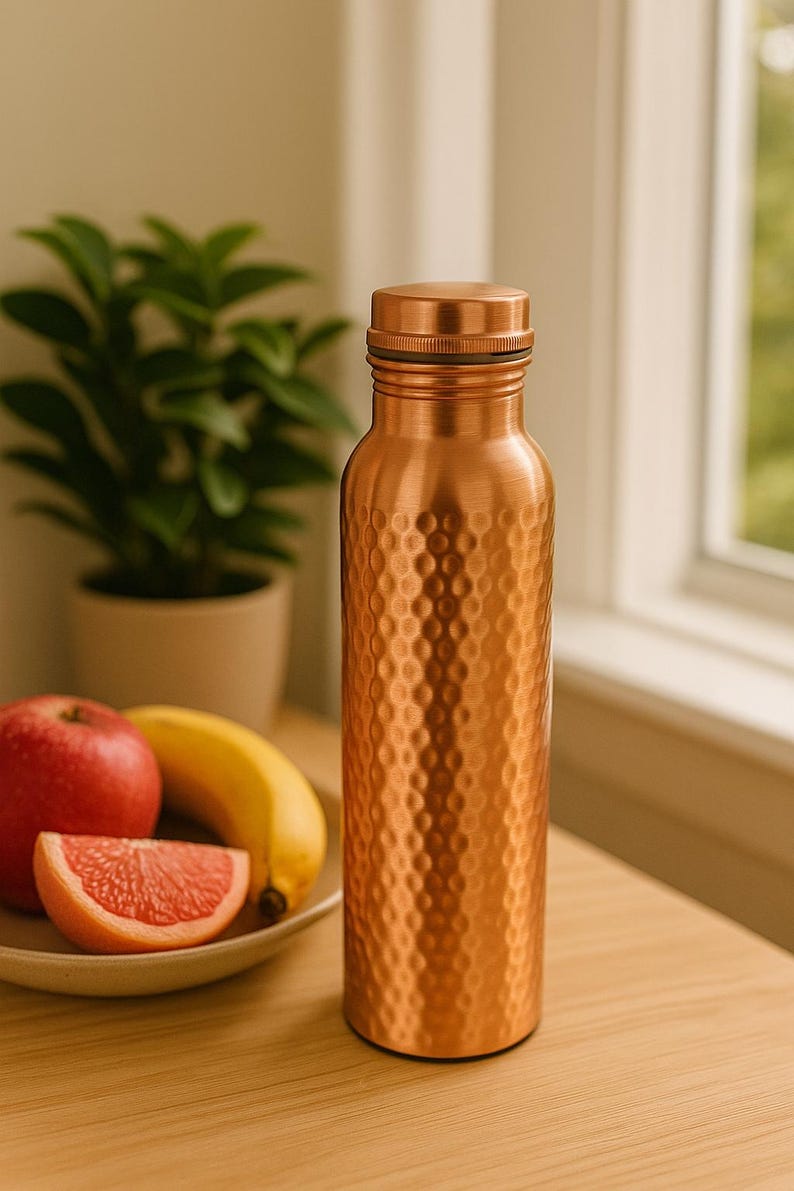 May include: A copper-colored water bottle with a textured, dimpled surface. The bottle is cylindrical with a screw-on cap. A red apple, a banana, and a grapefruit slice are on a plate next to the bottle. A potted plant is in the background.