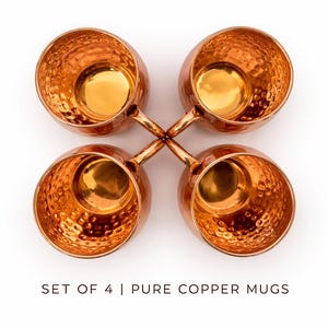 May include: Four copper mugs with a hammered texture, arranged in a cross shape against a white background. The mugs have a shiny, golden interior and small handles. The text at the bottom reads "SET OF 4 | PURE COPPER MUGS".