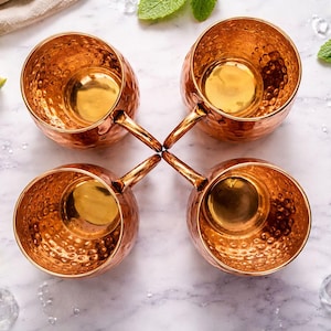 May include: Four copper Moscow Mule mugs with hammered exteriors are arranged in a square. Limes, mint sprigs, and ice cubes surround the mugs on a marble surface. The mugs have a capacity of approximately 16 ounces.