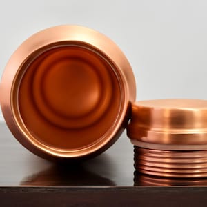 May include: A copper-colored water bottle with a matching lid. The bottle is open, revealing the interior. The lid is off to the side, showing the threaded design. The bottle and lid are made of a shiny metal.