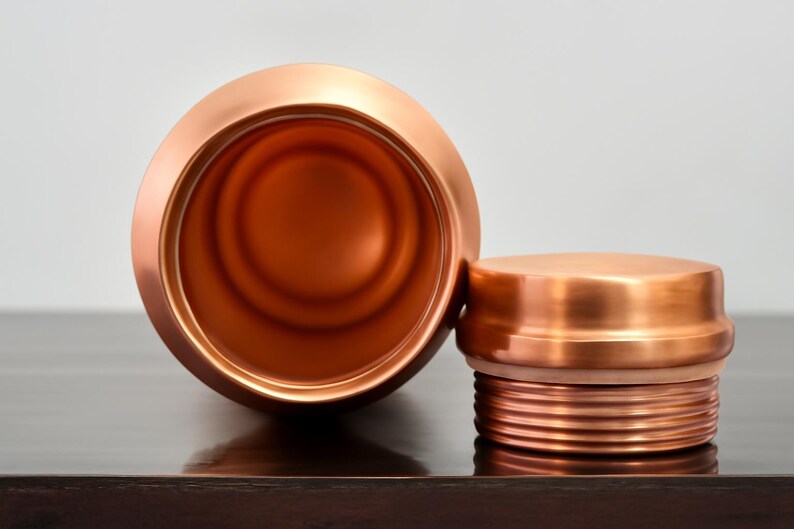 May include: A copper-colored water bottle with a matching lid. The bottle is open, revealing the interior. The lid is off to the side, showing the threaded design. The bottle and lid are made of a shiny metal.