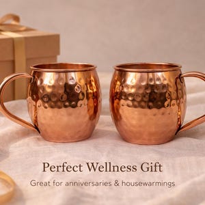 May include: Two copper Moscow Mule mugs with hammered texture and curved handles. A gift box with a gold ribbon is in the background. The text reads "Perfect Wellness Gift" and "Great for anniversaries & housewarmings".