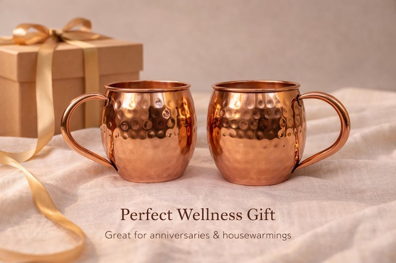 May include: Two copper Moscow Mule mugs with hammered texture and curved handles. A gift box with a gold ribbon is in the background. The text reads "Perfect Wellness Gift" and "Great for anniversaries & housewarmings".