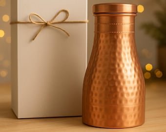 May include: A copper-colored water bottle with a hammered texture, next to a white gift box tied with twine. The bottle has a rounded shape and a screw-on lid. The gift box is rectangular.