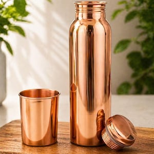 May include: A copper water bottle with a matching cup and lid. The bottle is tall and cylindrical, with a screw-on lid. The cup is short and round. All items have a shiny, metallic finish and sit on a wooden surface.