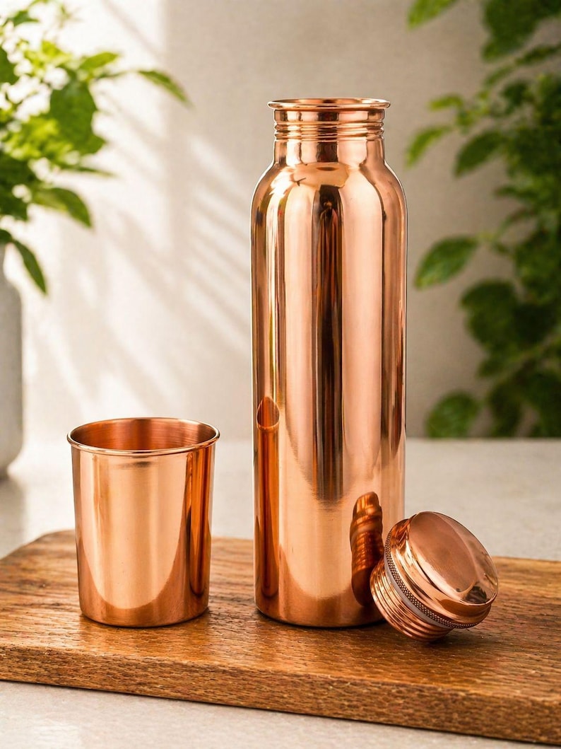 May include: A copper water bottle with a matching cup and lid. The bottle is tall and cylindrical, with a screw-on lid. The cup is short and round. All items have a shiny, metallic finish and sit on a wooden surface.