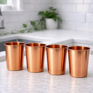 May include: Four copper-colored metal cups are arranged in a row on a light-colored countertop. The cups have a cylindrical shape with a slightly flared rim. The kitchen setting includes a window and a white tiled backsplash.