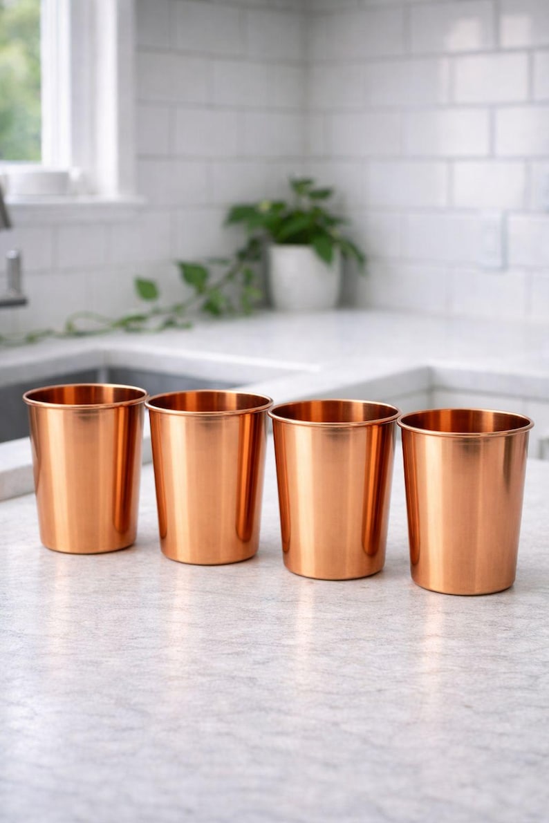May include: Four copper-colored metal cups are arranged in a row on a light-colored countertop. The cups have a cylindrical shape with a slightly flared rim. The kitchen setting includes a window and a white tiled backsplash.