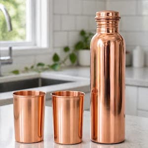 May include: A copper water bottle and two matching copper cups sit on a countertop. The bottle has a screw-on cap. The items are made of shiny copper and reflect the light. The kitchen background includes a window and white tile.