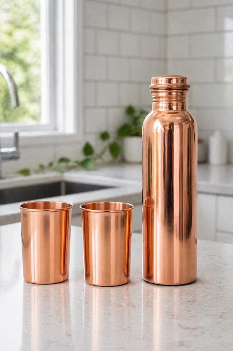 May include: A copper water bottle and two matching copper cups sit on a countertop. The bottle has a screw-on cap. The items are made of shiny copper and reflect the light. The kitchen background includes a window and white tile.