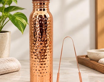 Puede incluir: Una botella de agua cilíndrica de cobre con textura martillada y tapa de rosca a juego, acompañada de un raspador de lengua de cobre fino en forma de U con agarres texturizados en los extremos, ambos con un acabado metálico pulido.