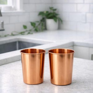 May include: Two copper-colored metal tumblers sit side-by-side on a light gray countertop. The cylindrical cups have a shiny, reflective surface and a simple, modern design. The kitchen setting includes a white tiled backsplash and a window.