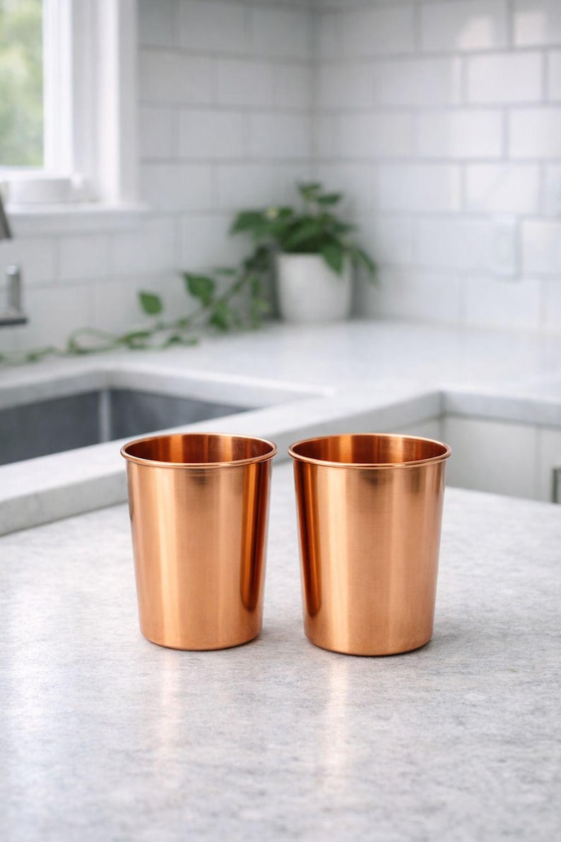 May include: Two copper-colored metal tumblers sit side-by-side on a light gray countertop. The cylindrical cups have a shiny, reflective surface and a simple, modern design. The kitchen setting includes a white tiled backsplash and a window.