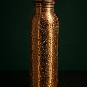 Könnte beinhalten: Eine kupferfarbene Wasserflasche mit strukturierter Oberfläche und Schraubverschluss. Die Flasche ist mit Wassertropfen bedeckt, was darauf hindeutet, dass sie kalt ist. Der Hintergrund ist dunkelgrün.