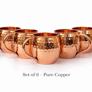 May include: Set of six copper mugs with a hammered finish. Each mug has a rounded body, a curved handle, and a shiny, reflective surface. The mugs are arranged in a semi-circle on a white background. Text at the bottom reads "Set of 6 - Pure Copper."