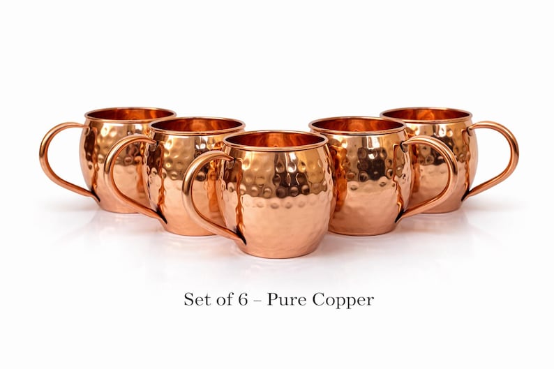 May include: Set of six copper mugs with a hammered finish. Each mug has a rounded body, a curved handle, and a shiny, reflective surface. The mugs are arranged in a semi-circle on a white background. Text at the bottom reads "Set of 6 - Pure Copper."