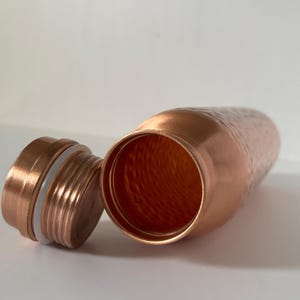 Puede incluir: Una botella de agua de color cobre con una superficie texturizada y una tapa de rosca a juego. La tapa est&aacute; quitada, revelando la abertura de la botella. La botella est&aacute; dise&ntilde;ada para contener l&iacute;quidos y es de metal.