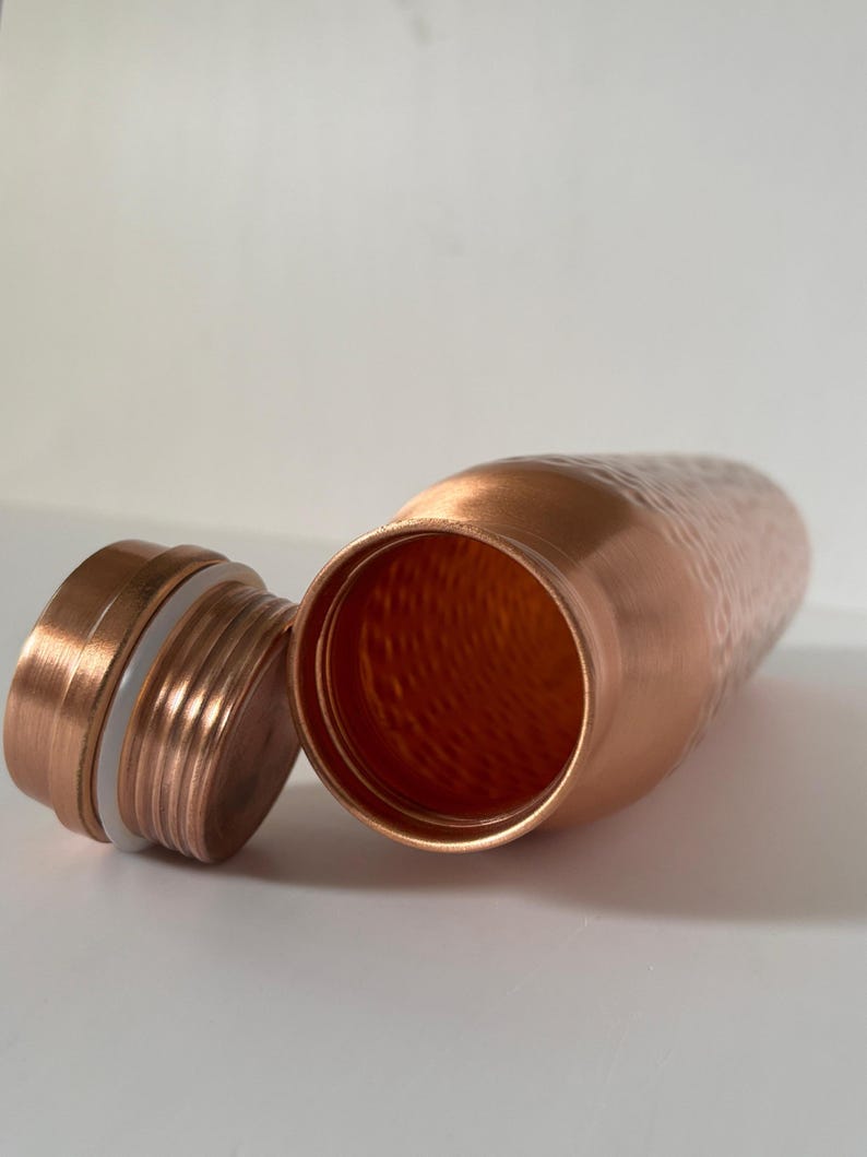 Puede incluir: Una botella de agua de color cobre con una superficie texturizada y una tapa de rosca a juego. La tapa est&aacute; quitada, revelando la abertura de la botella. La botella est&aacute; dise&ntilde;ada para contener l&iacute;quidos y es de metal.