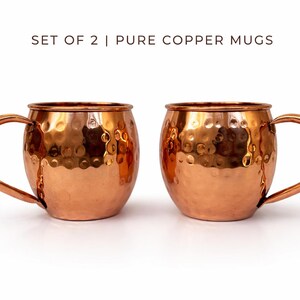 May include: Two copper mugs with a hammered finish and curved handles. The mugs are a shiny copper color and are set against a white background. The text "SET OF 2 | PURE COPPER MUGS" is at the top.