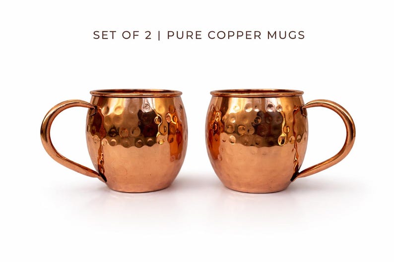 May include: Two copper mugs with a hammered finish and curved handles. The mugs are a shiny copper color and are set against a white background. The text "SET OF 2 | PURE COPPER MUGS" is at the top.