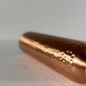 Puede incluir: Un objeto cil&iacute;ndrico de color cobre con una textura martillada. La superficie refleja la luz, creando reflejos y sombras. El objeto parece ser un recipiente o un elemento decorativo.