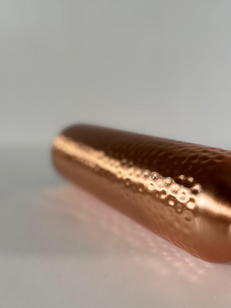 Puede incluir: Un objeto cil&iacute;ndrico de color cobre con una textura martillada. La superficie refleja la luz, creando reflejos y sombras. El objeto parece ser un recipiente o un elemento decorativo.