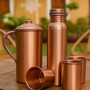 Pode incluir: Um conjunto de utensílios para beber de cor cobre, incluindo um jarro com tampa, uma garrafa de água e três pequenas chávenas. Os itens estão dispostos sobre uma superfície de madeira, refletindo um brilho metálico quente. O fundo está desfocado.