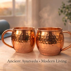 May include: Two copper mugs with a hammered texture, each with a curved handle. Steam rises from the mugs, suggesting a hot beverage. The text "Ancient Ayurveda &bull; Modern Living" is visible.