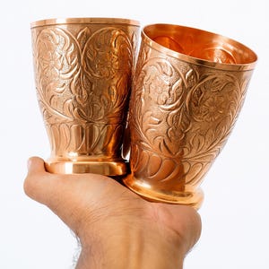 May include: Two copper-coloured metal drinking glasses with detailed floral and leaf patterns. The glasses have a wide base and a slightly flared rim. The glasses are held in a hand against a white background.