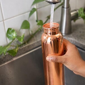 May include: A copper water bottle being filled with water from a stainless steel faucet. The bottle is cylindrical with a narrow neck. The background includes a stainless steel sink, white tile, and green leafy plants.